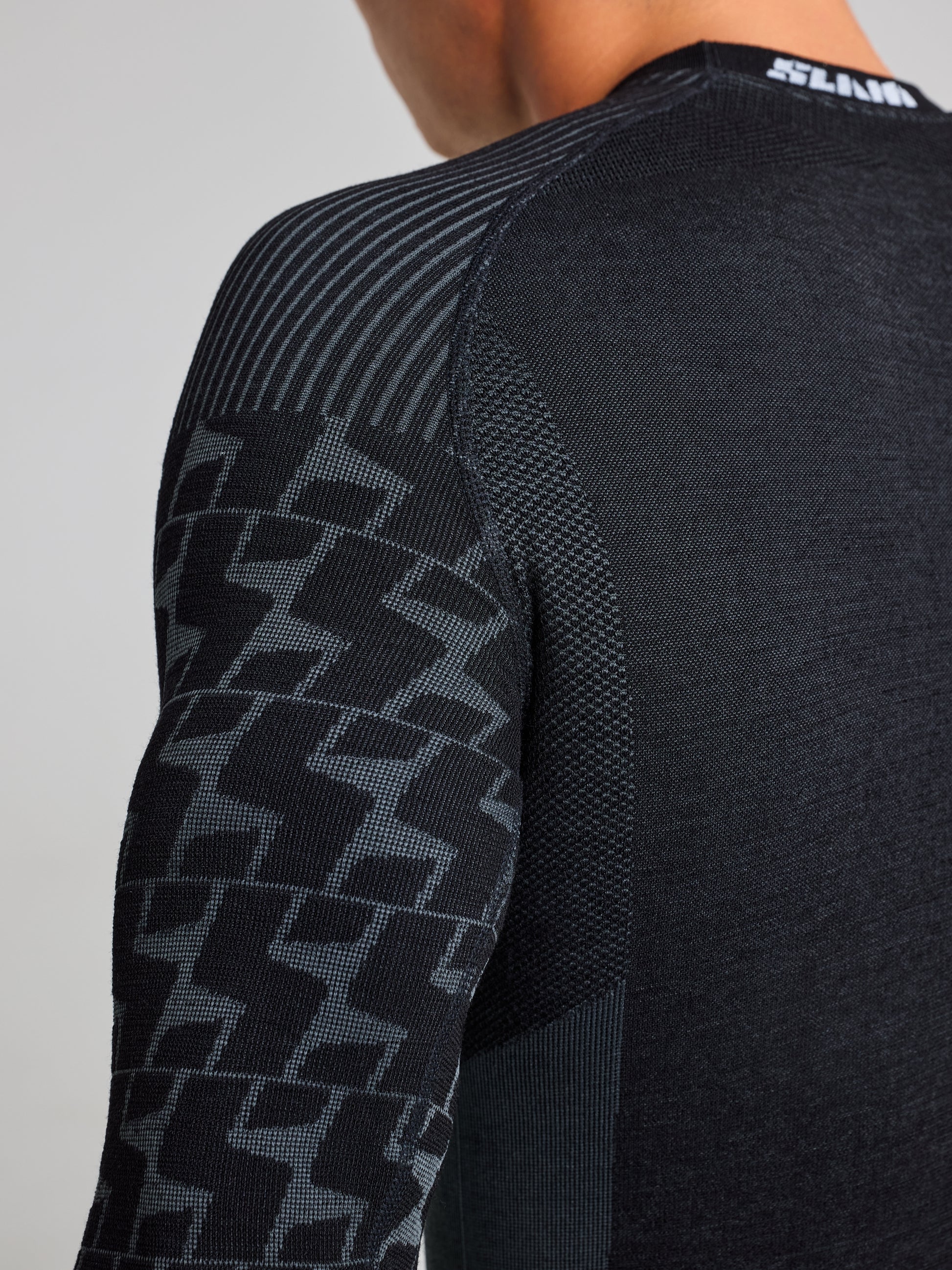 A person wearing the PRO SEAMLESS MERINO TOP by SLAM, featuring a fitted black athletic design with textured patterns and zigzag designs on the sleeves. Its quick-drying, ergonomic design ensures comfort, while a partially visible brand logo is located at the back of the neck.