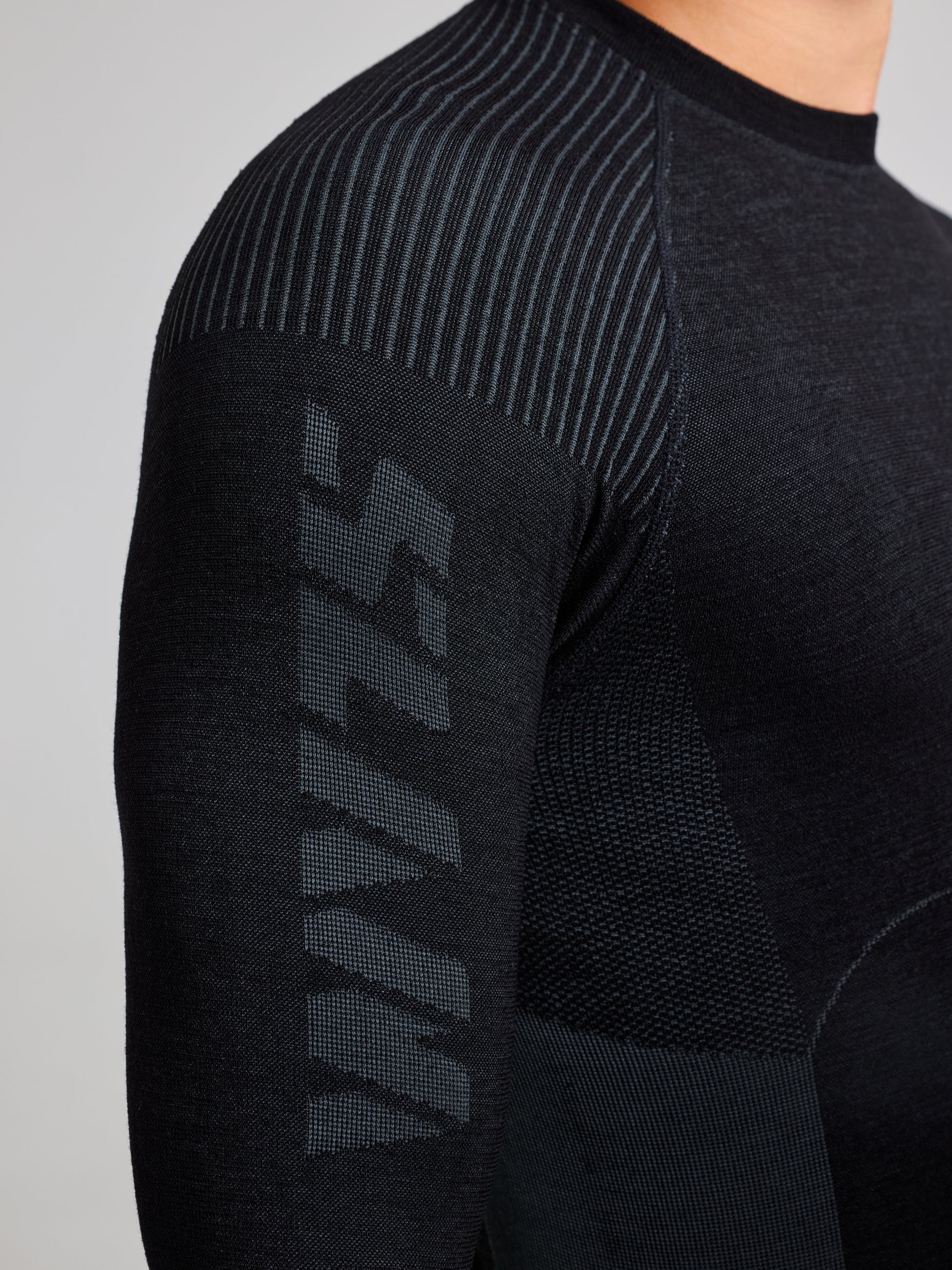 Close-up of a person wearing the SLAM PRO SEAMLESS MERINO TOP in black. The fabric features ribbed detailing and a textured pattern for an ergonomic fit. The sleeve displays the word "Vavts" in stylized letters, highlighting the garment's modern design.