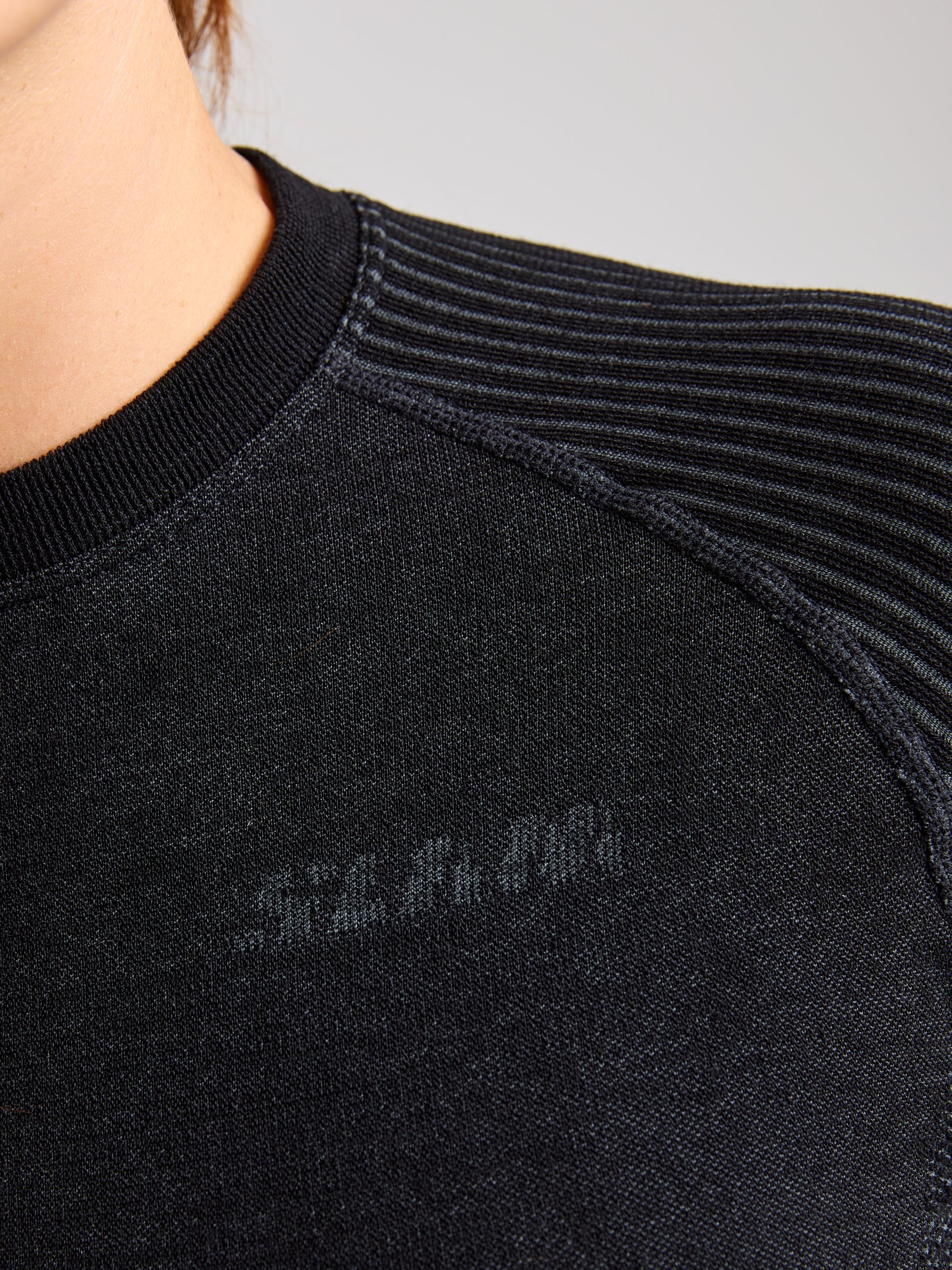 Close-up of a person wearing SLAM's PRO SEAMLESS MERINO TOP in black. The focus is on the ribbed, textured fabric featuring a subtle pattern near the shoulder. The ergonomic design enhances comfort, while their face and hair are partially visible in the top left corner.