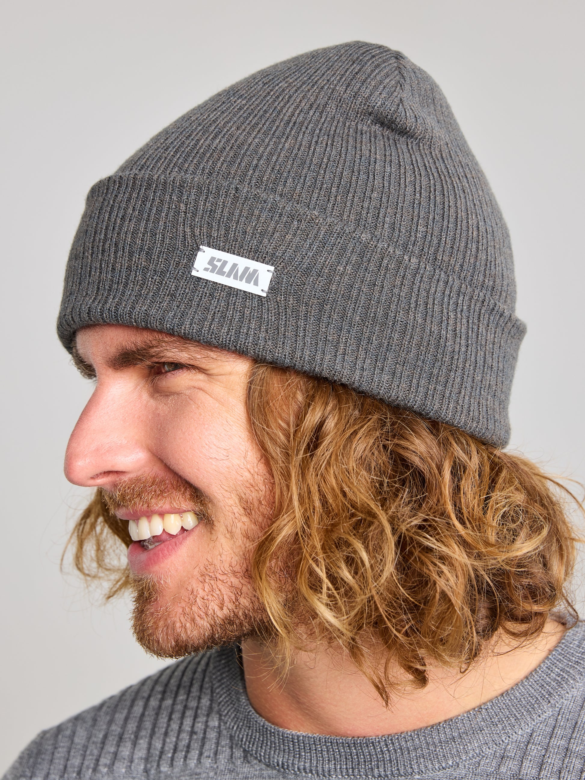 A person with long, wavy hair is wearing a SLAM CABLE BEANIE in gray and a sweater. Made from recycled yarn, the beanie includes a small tag on the side. The unisex winter accessory enhances their appearance as they smile looking to the right.