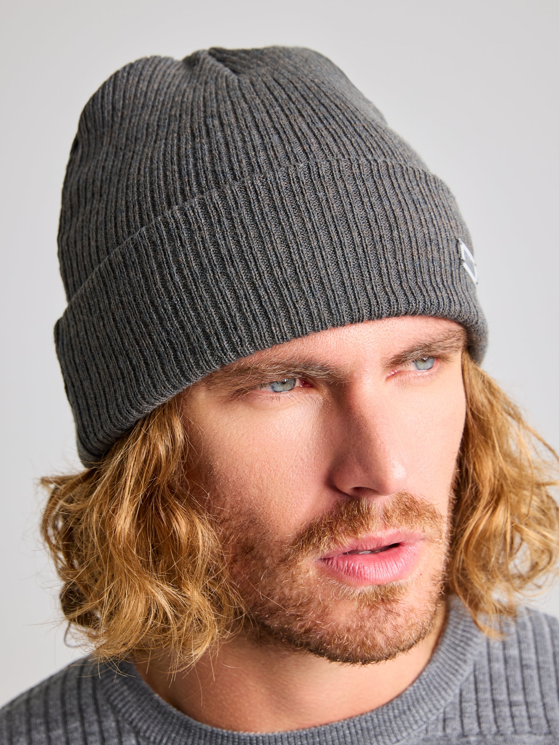 A person with long, wavy blond hair and a beard is seen wearing the SLAM CABLE BEANIE, made from recycled yarn in a gray hue, along with a complementary sweater. The backdrop features a simple, light tone.