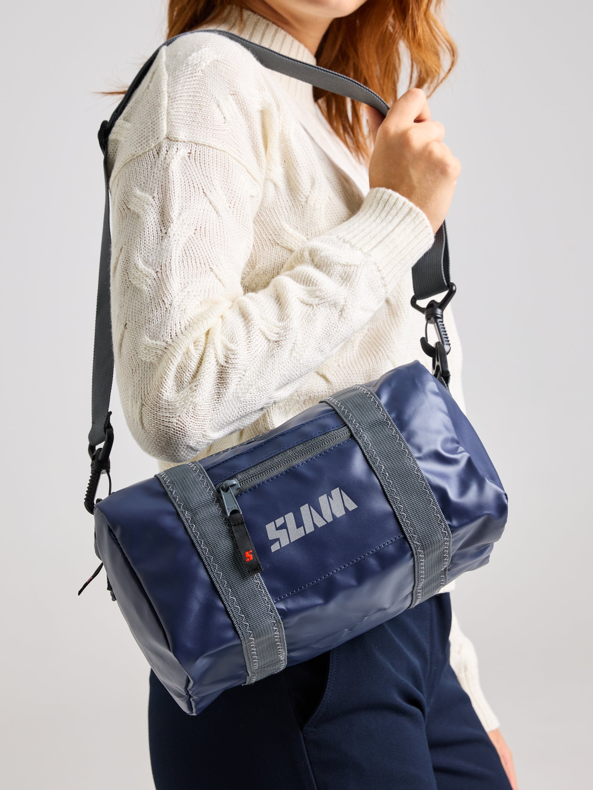 A person wearing a white sweater and dark pants holds an XS-sized blue cylindrical duffle bag by SLAM, featuring a black shoulder strap. The water-repellent WR Duffle Bag comes with the brand name "SLAVA" on it and includes a zippered front pocket.