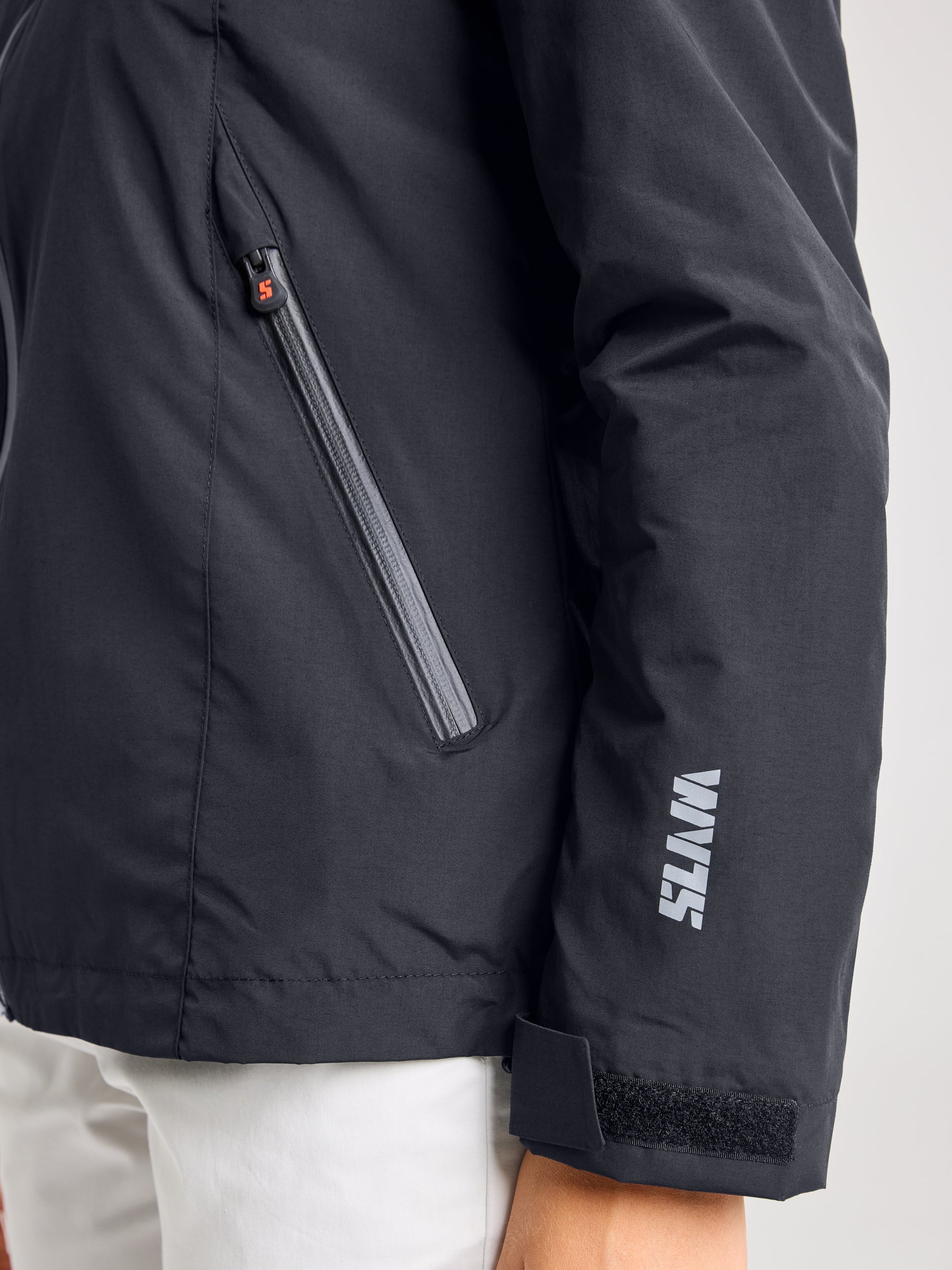 A close-up of a person wearing the SLAM CREW WOMAN WARM HOODED JACKET in black, showcasing its zippered pocket and Velcro-adjusted cuff—ideal for navigating the seas. The sleeve features "WATTS" in white lettering. Made from waterproof material, it complements light-colored pants with style.