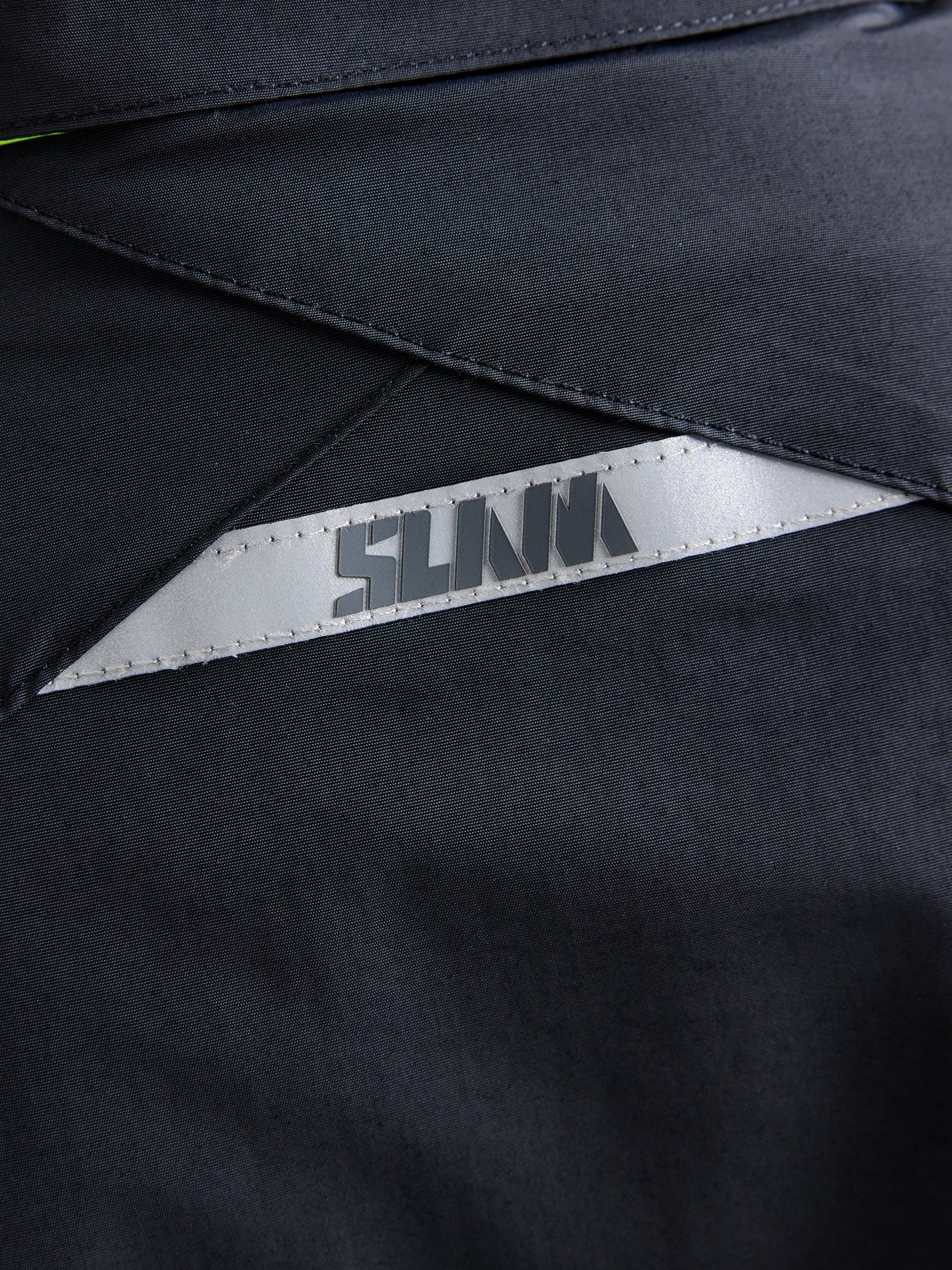 Close-up of the black fabric from the CREW WOMAN WARM HOODED JAKET by SLAM, showcasing a triangular, metallic patch with "SLAVA" printed on it. The visible stitching and texture emphasize the high-quality polar fleece material.