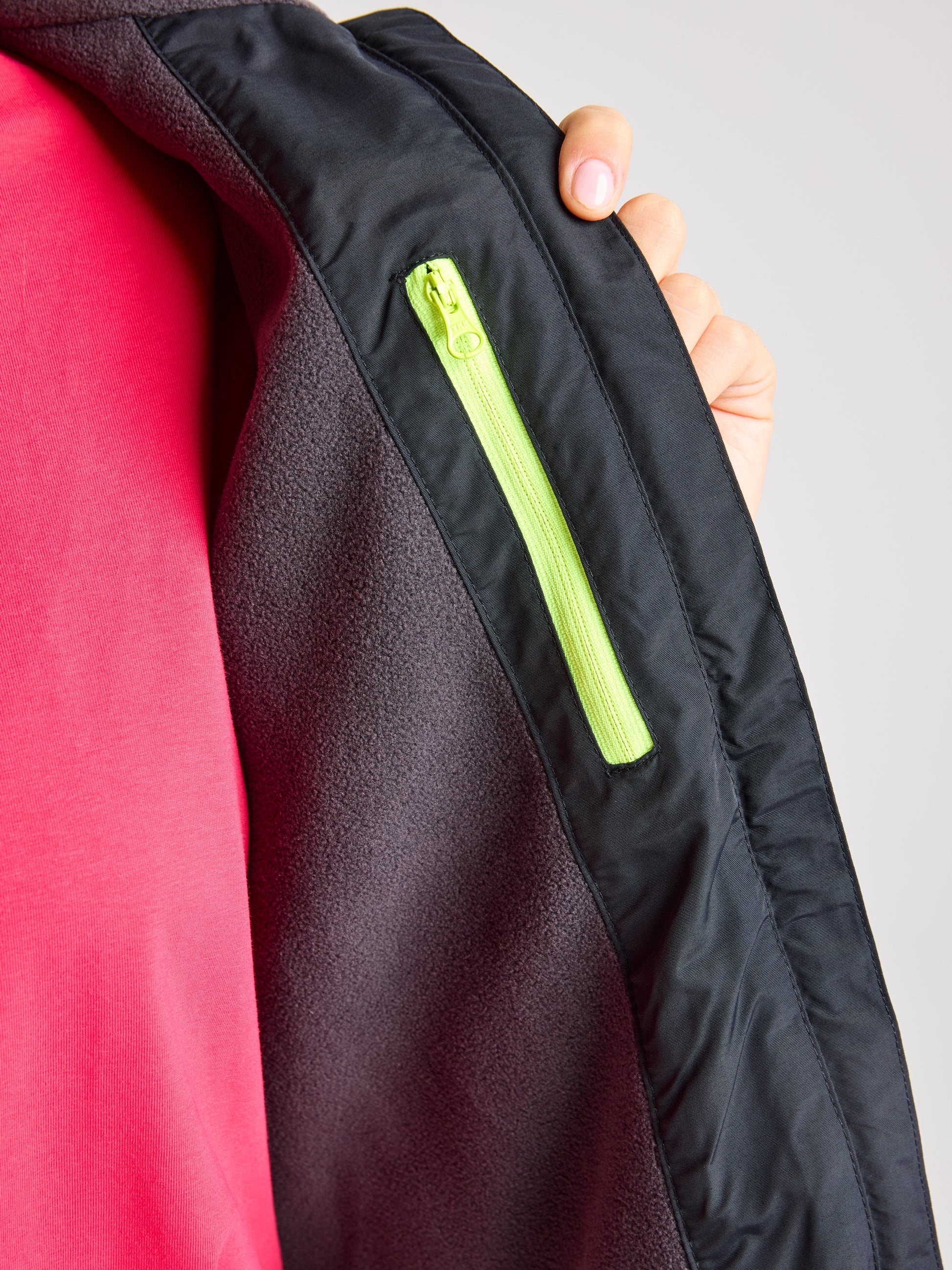 A person is holding open a SLAM CREW WOMAN WARM HOODED JACKET, showcasing the polar fleece inner lining. The interior includes a zippered pocket with a bright green zipper. The person's thumb is visible, and they are wearing a red shirt.