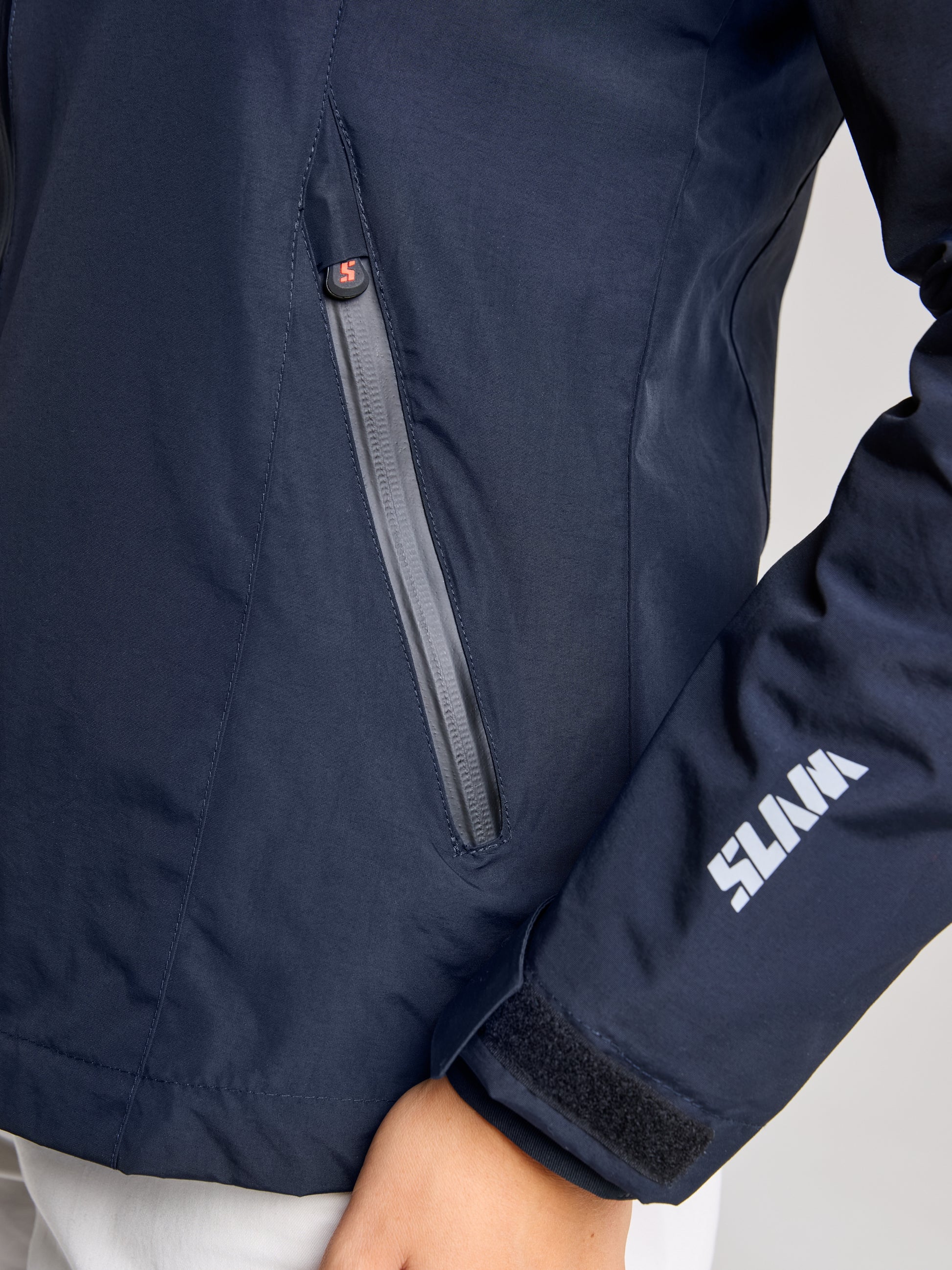 A close-up of a person wearing the navy CREW WOMAN WARM HOODED JAKET by SLAM. The left sleeve displays a white logo, and there's a zippered pocket on the side. This waterproof jacket has a smooth texture, with the person standing and their arm slightly bent.