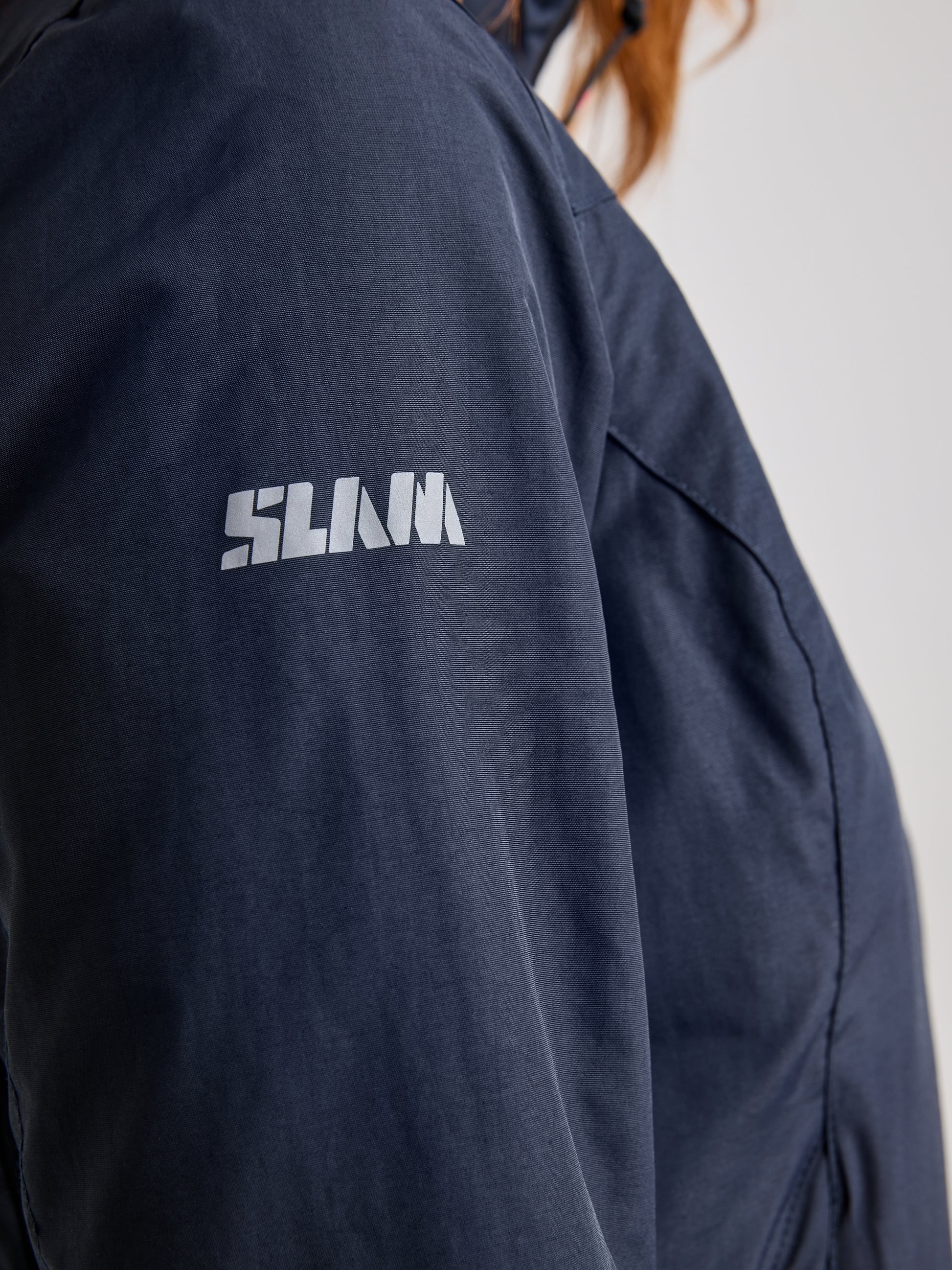 Close-up view of a person wearing a dark blue CREW WOMAN WARM HOODED JACKET by SLAM, featuring "SLAM" printed in white on the sleeve. The jacket's waterproof design stands out against the blurred background.