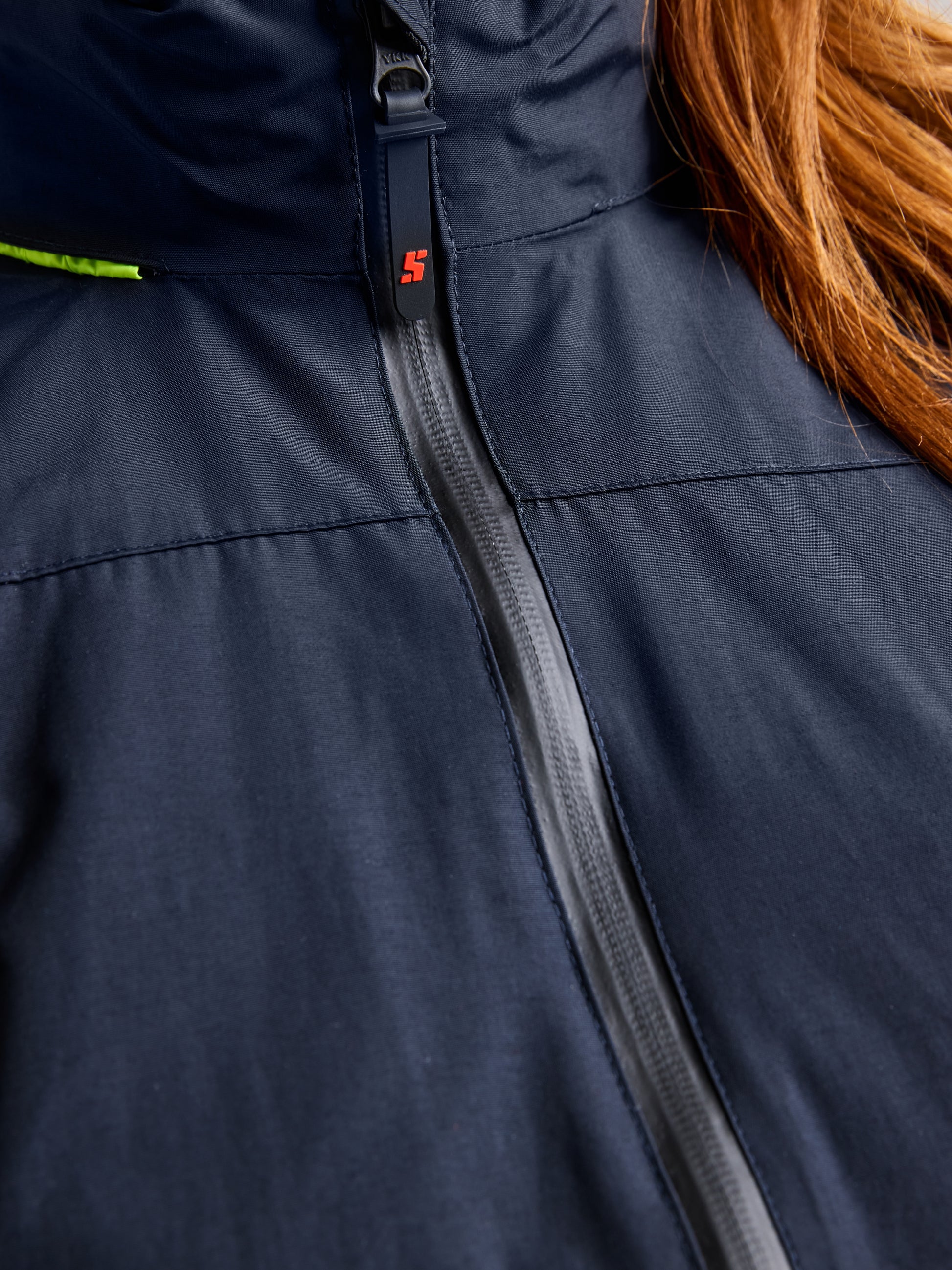 Close-up of the upper section of the SLAM CREW WOMAN WARM HOODED JAKET in navy blue, showcasing a zipper adorned with a small red logo. A glimpse of long, brown hair on the right side emphasizes its waterproof design and snug polar fleece lining.