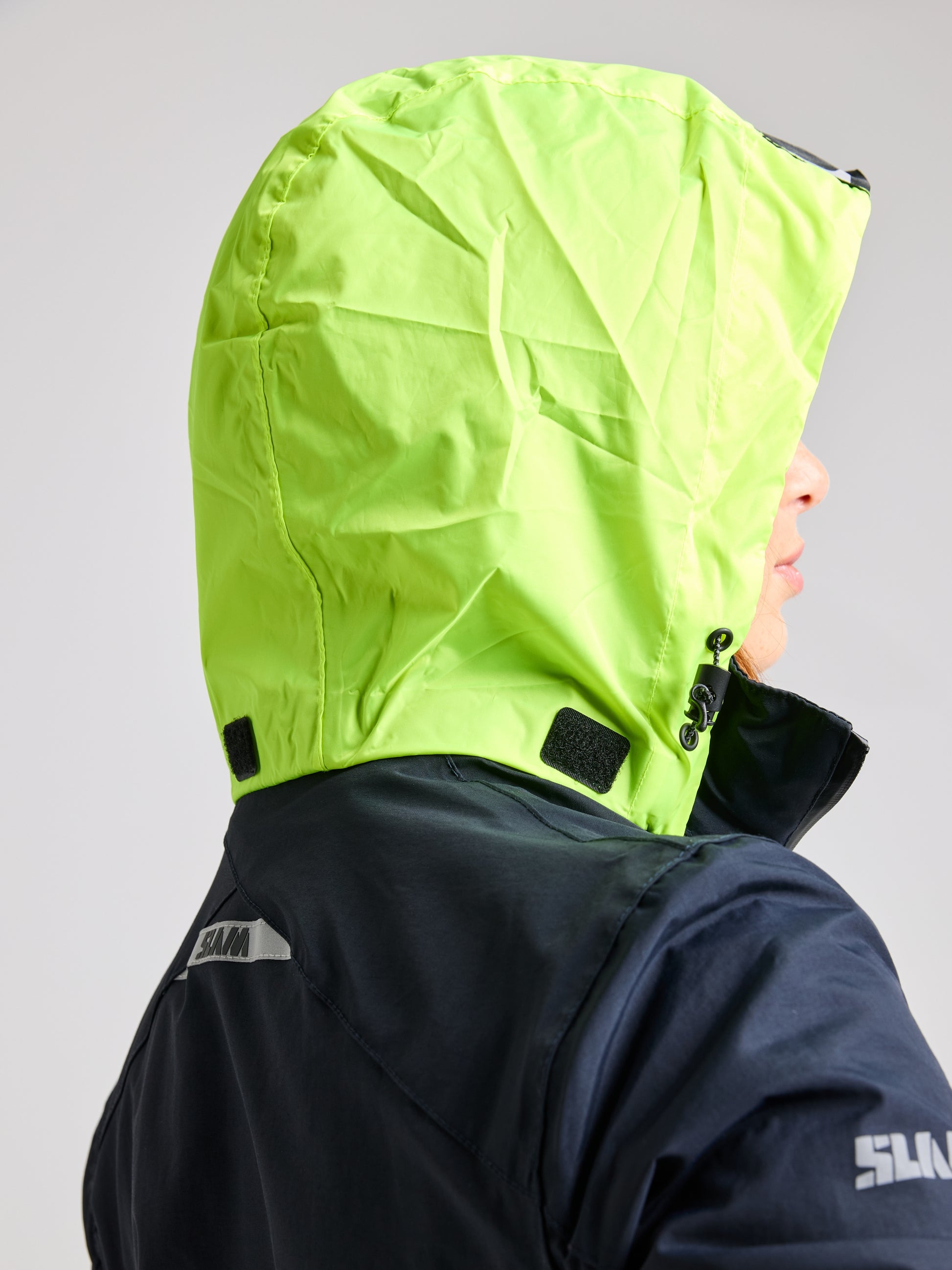 A person is wearing the SLAM CREW WOMAN WARM HOODED JACKET in bright green, partially turned away from the camera. This jacket, designed with waterproof material, displays a black and white logo on the back. The hood is fastened with black Velcro patches.