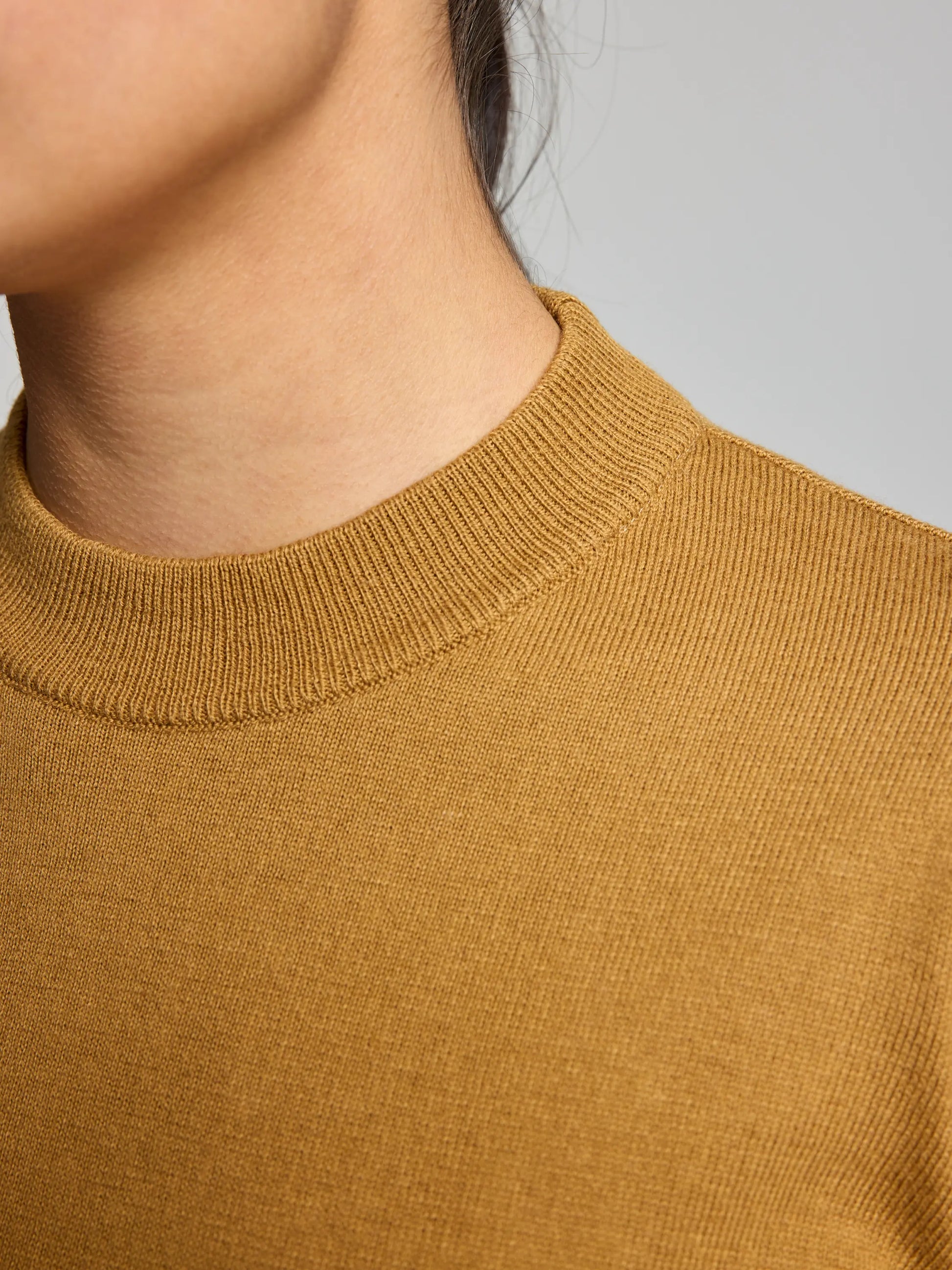 A close-up image features a person wearing the WOMAN JUMPER YONNE CL by SLAM, a mustard-colored ribbed sweater made from a cotton/wool blend. The emphasis is on the texture and mock neckline of this women's crew-neck style, with the person's face partially out of frame.