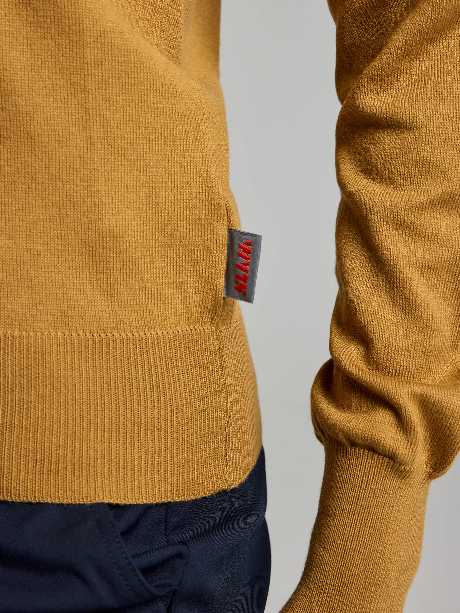 A close-up image shows someone wearing the WOMAN JUMPER YONNE CL by SLAM, a mustard-colored sweater made from a soft cotton/wool blend with a crew neck. A distinctive red and gray tag is visible on the side. The sweater is perfectly paired with dark blue pants, and the wearer’s right arm is slightly bent at the elbow.