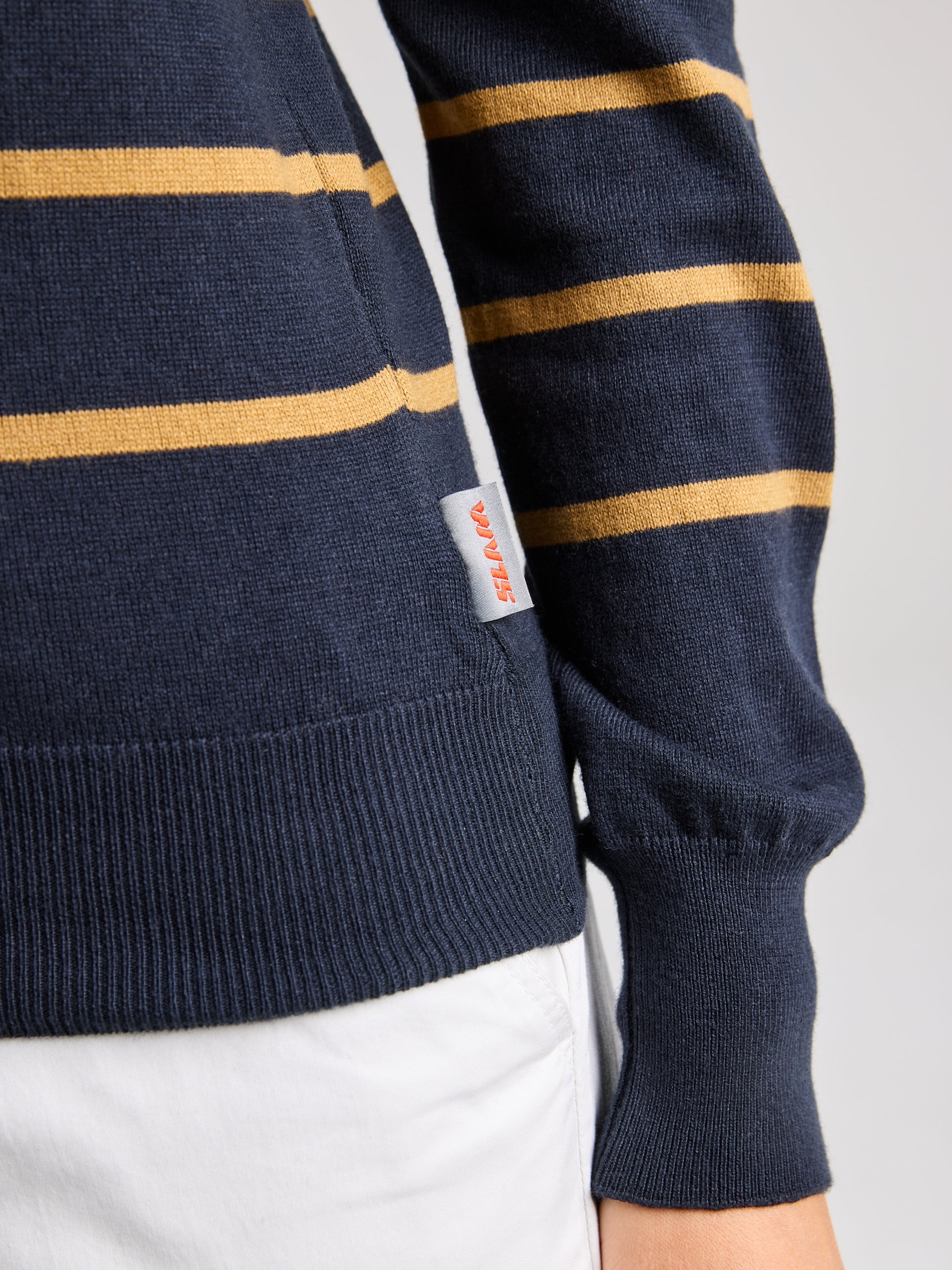 A close-up shows a person wearing the WOMAN JUMPER YONNE CL from SLAM, a women's crew-neck sweater made from a cotton/wool blend. The design features dark blue with mustard-colored stripes, complemented by ribbed cuffs and a small tag with text. It pairs perfectly with white pants.