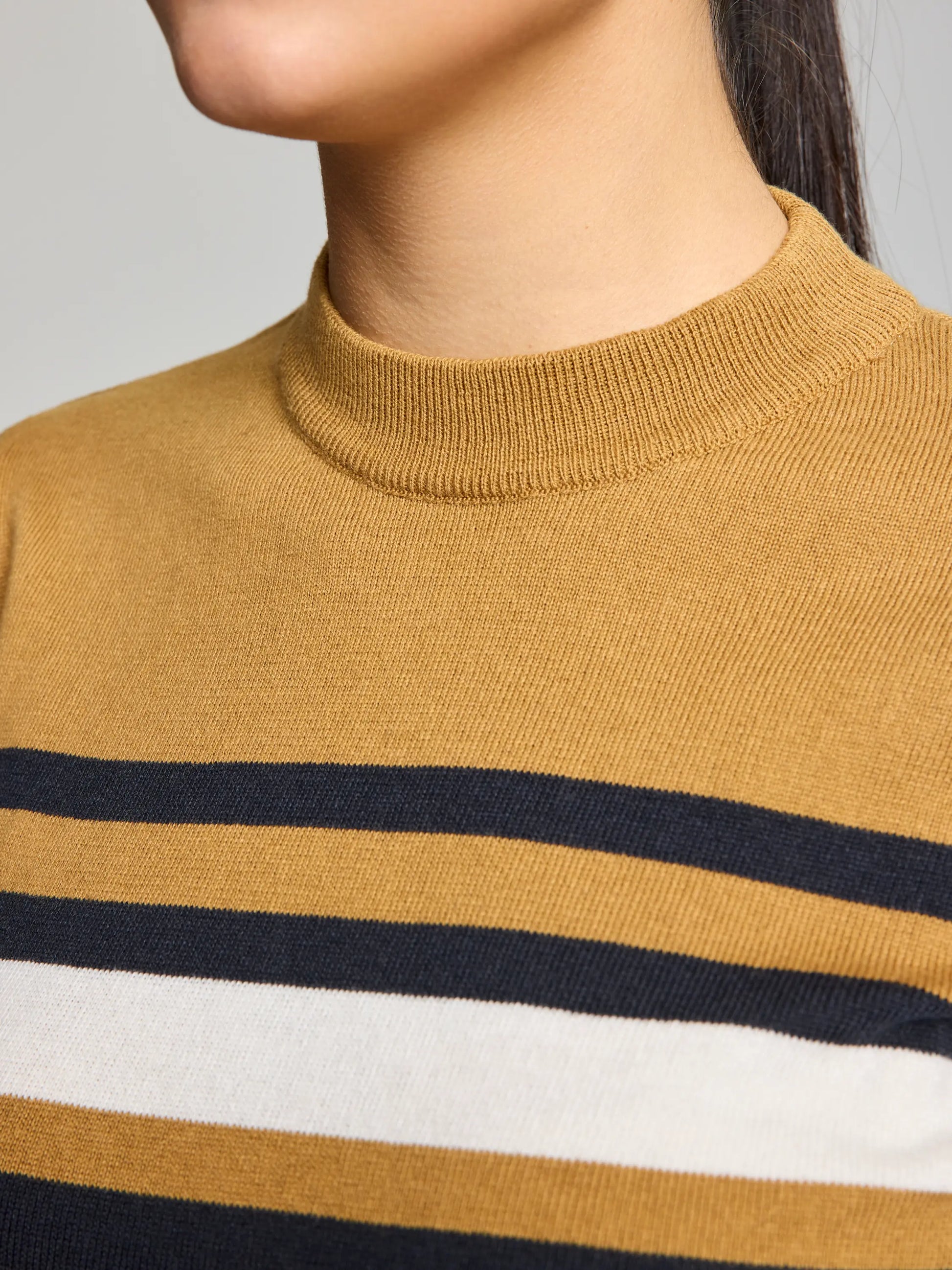 A close-up shows someone wearing the WOMAN JUMPER YONNE CL by SLAM. This women's crew-neck sweater comes in mustard with a high neckline and is made from a cozy cotton/wool blend. It features stylish horizontal stripes in black, white, and mustard colors. The person's face is partially visible from the side.