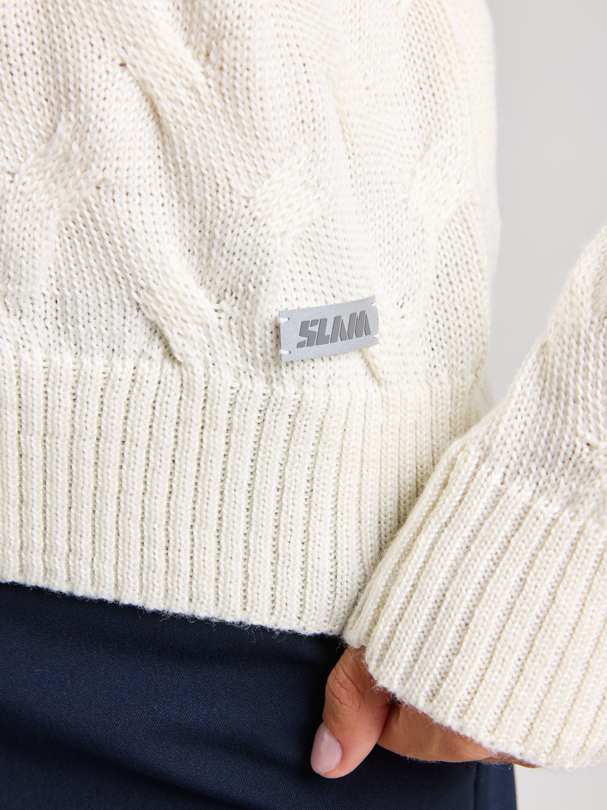 A close-up shows a person wearing the SEAWOOL WOMAN CABLE JUMPER, a cream-colored crewneck sweater made from Merino-blend yarn, featuring a gray "SLAM" tag near the hem. The person's hand is partially visible, and they are paired with dark pants, highlighting the texture of the sweater.