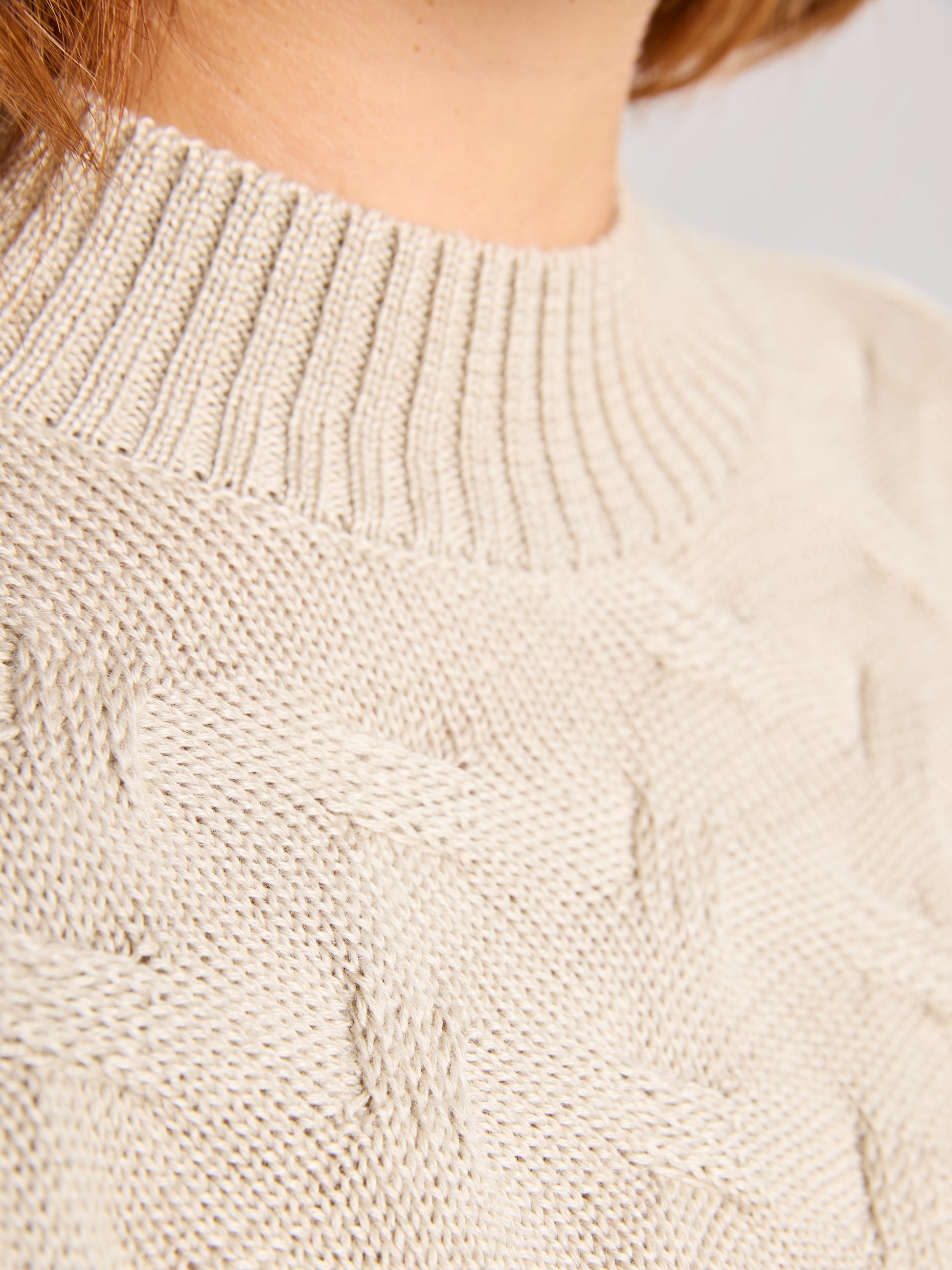 A close-up of the SEAWOOL WOMAN CABLE JUMPER by SLAM highlights the textured knitting pattern and ribbed crew neckline in beige Merino-blend yarn. The neutral, blurred background accentuates the jumper's intricate design.