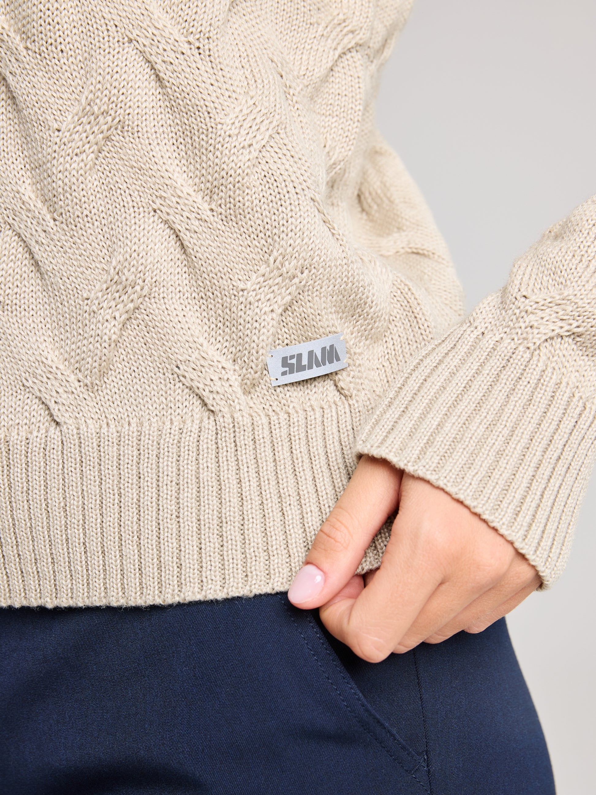 A person wearing a SEAWOOL WOMAN CABLE JUMPER in beige, adorned with a metallic tag that displays the brand name SLAM. Made from Merino-blend yarn, this jumper seamlessly combines style and comfort. They pair it with dark blue pants and gently hold the hem of the sweater between their thumb and index finger.