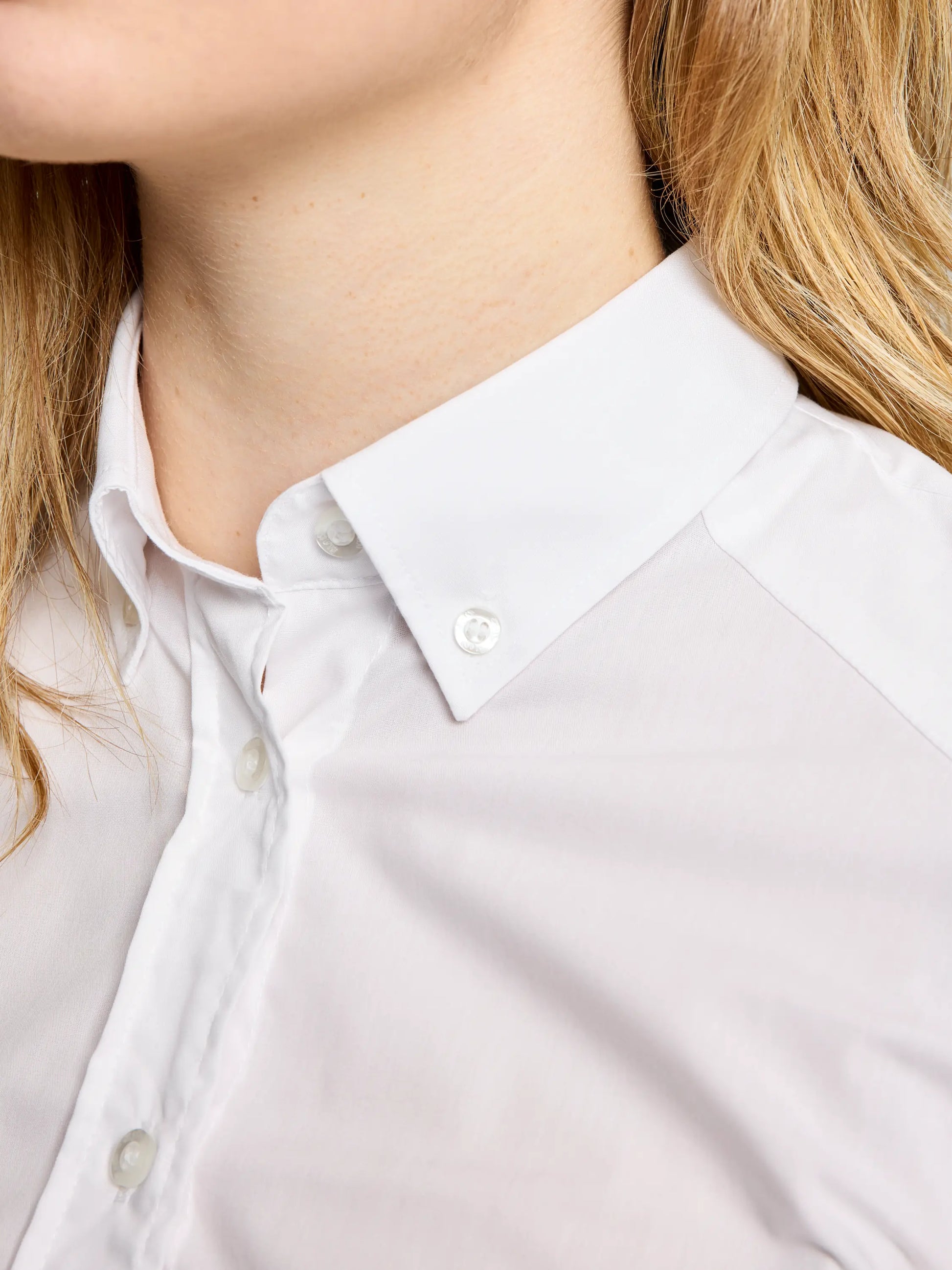 Close-up of a person wearing the DECK WOMAN NO IRON SHIRT CL from SLAM, featuring a classic cut in pure white cotton. The focus is on the crisp collar and top buttons, with their wavy blonde hair partially visible on the left side.