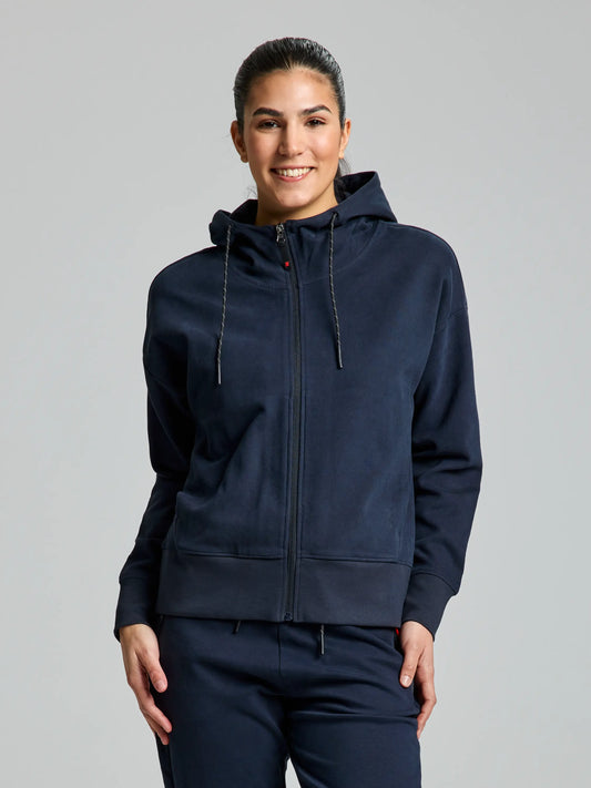 A person wearing the DECK WOMAN FULL ZIP HOODIE from SLAM in dark navy, along with matching sweatpants, stands against a plain gray background. This warm cotton hoodie, ideal for sporting activities, is zipped up as the person places their hands in the pockets. The hood is down, and they are smiling.