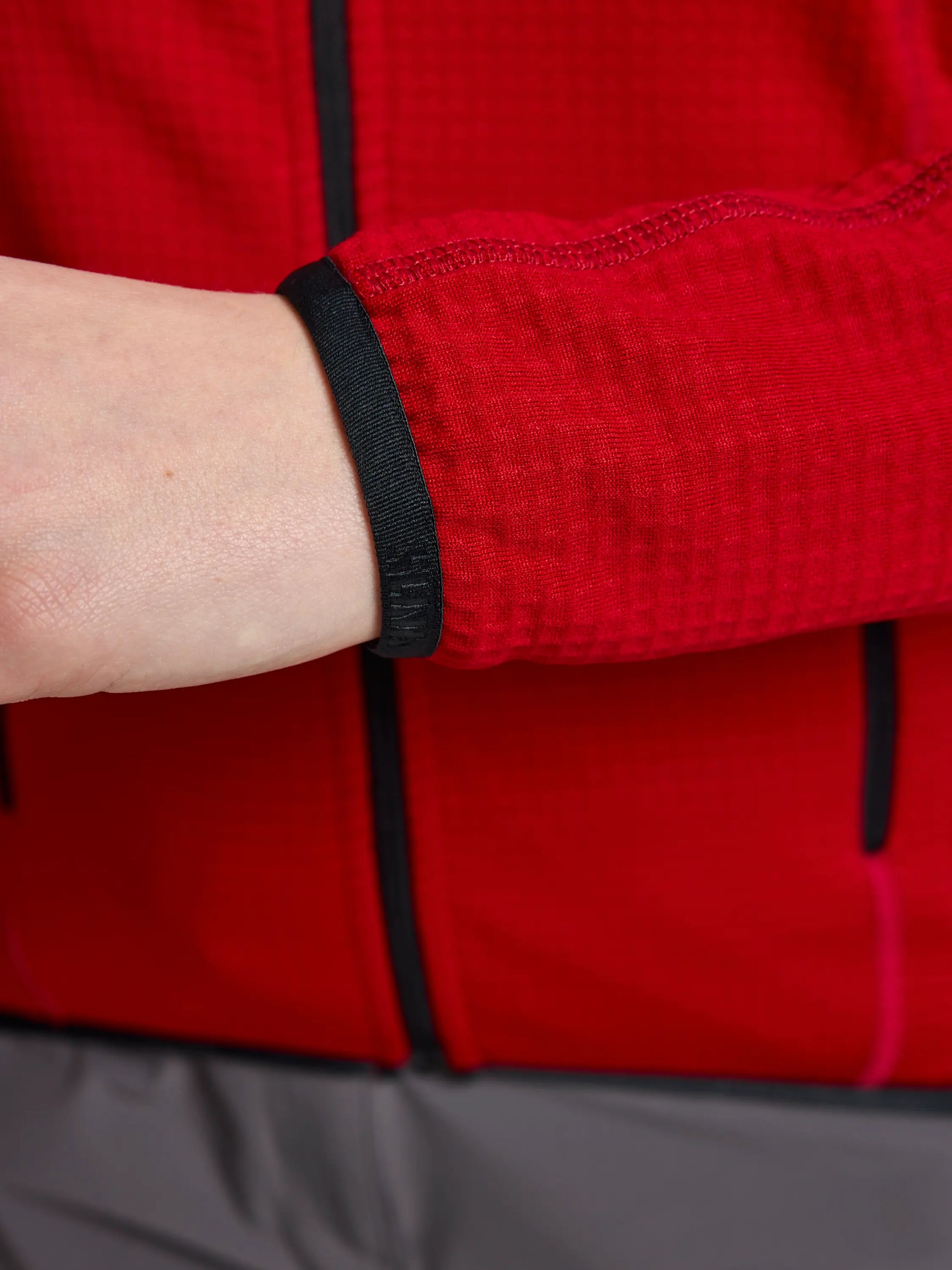 A close-up of a person wearing the ACTIVE WOMAN GRID FLEECE CL by SLAM, showcasing its red, long-sleeved design with Pontetorto's Grid Fleece and black elastic cuffs, paired with a hint of gray bottom garment. The textured checkered pattern, highlighted by black seams, provides breathable comfort. The person's hand is partially hidden.