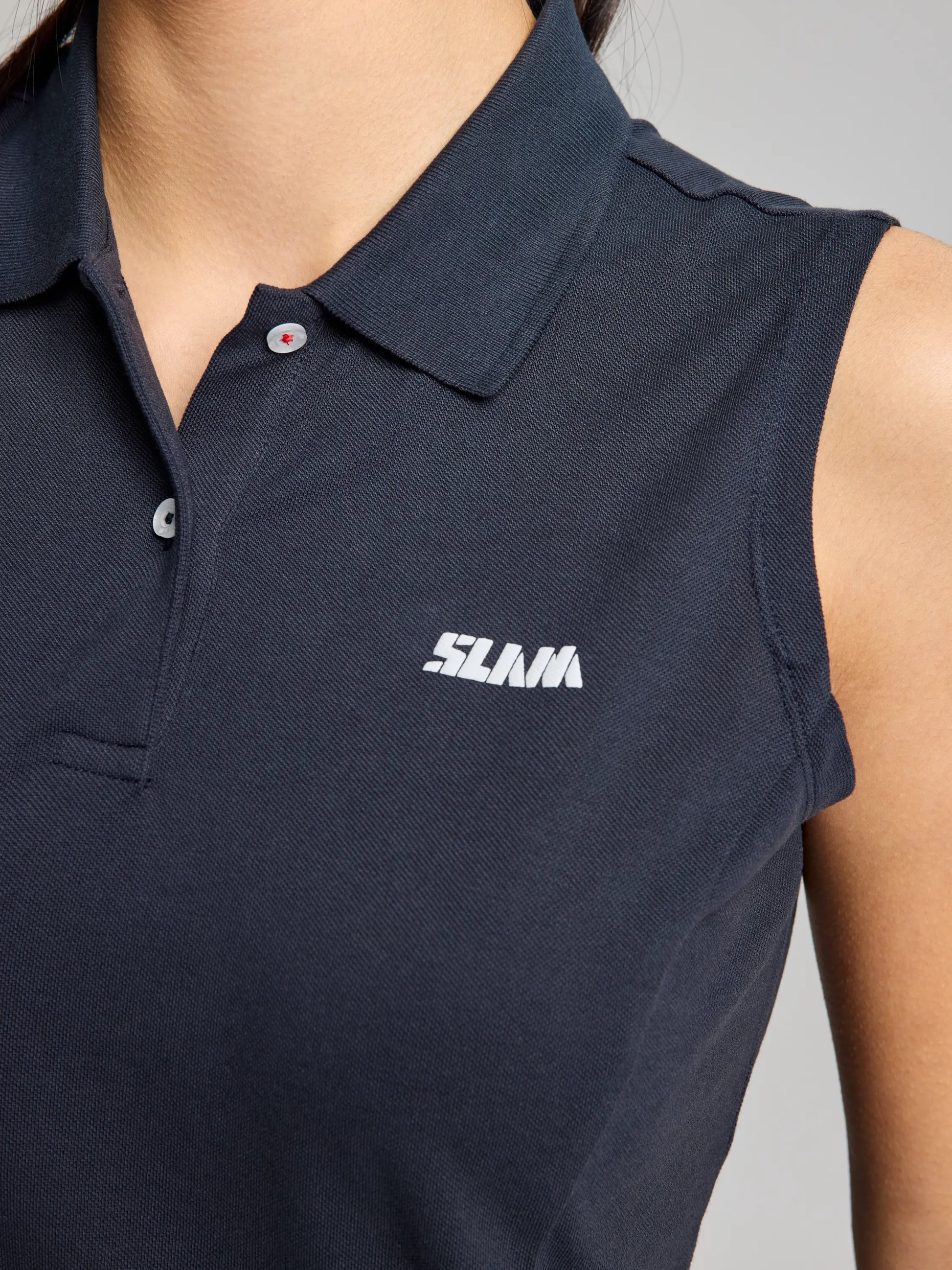 Close-up of the DOCK WOMAN POLO SLEEVELESS CL featuring a collar, crafted from pique cotton. This black polo shirt, branded by SLAM, is adorned with the word "SLAM" in white lettering on the left chest and includes buttons along with a visible weave texture.