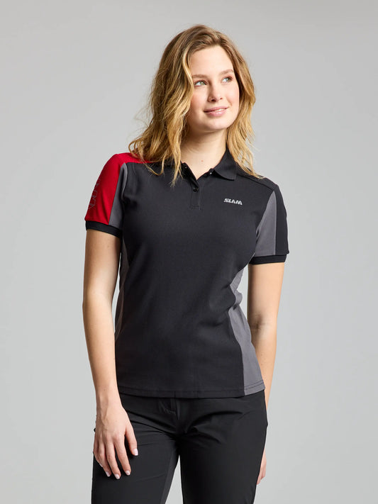 A person with wavy hair is wearing the WOMAN LOGO TACTEL POLO CL by SLAM, in black, red, and gray. Paired with black pants, the polo features a small logo on the chest and provides UFP 30+ protection. The backdrop is a plain gray.