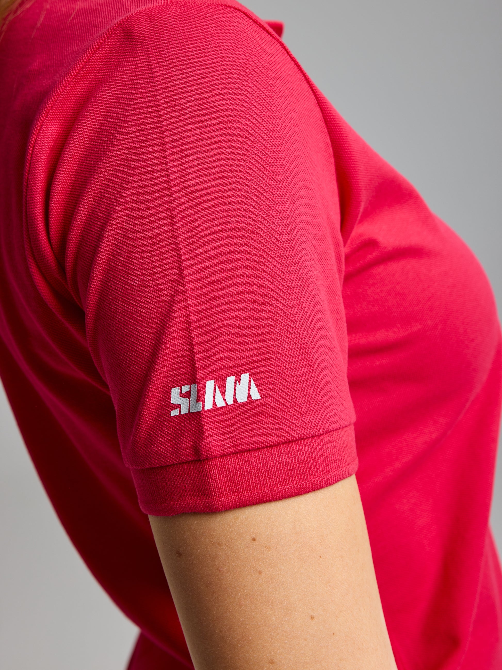 A person wearing a DECK WOMAN POLO SHORT SLEEVE in vibrant pink by SLAM. The soft cotton pique fabric and the small white "SLNA" sleeve logo contrast with the neutral background, enhancing the shirt's bold hue.