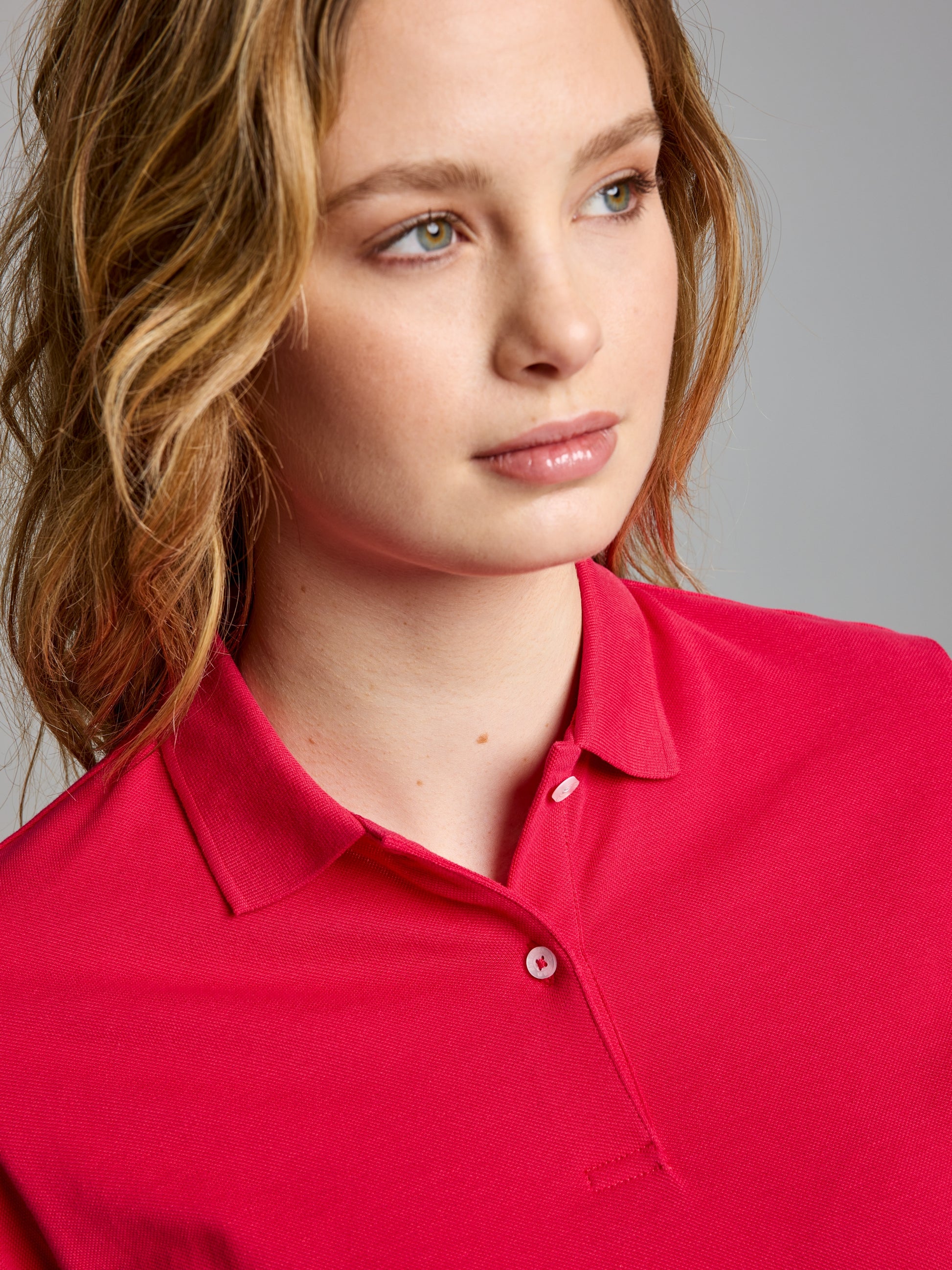 A person with shoulder-length hair gazes to the side, wearing a vibrant DECK WOMAN POLO SHORT SLEEVE by SLAM in bright pink cotton pique. The neutral gray background accentuates their striking attire.