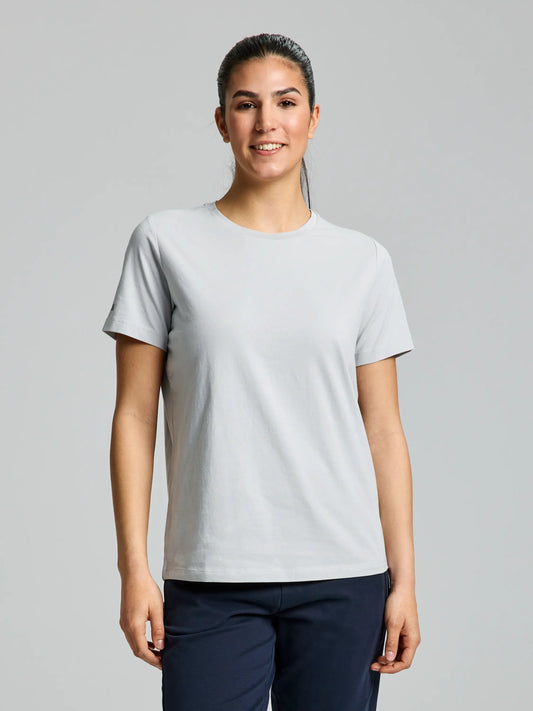 A person with long dark hair tied back is wearing a comfortable DECK WOMAN T SHIRT by SLAM in glacier grey and dark pants, standing against a plain gray background. Made from natural cotton, they have a neutral expression and their arms by their sides.