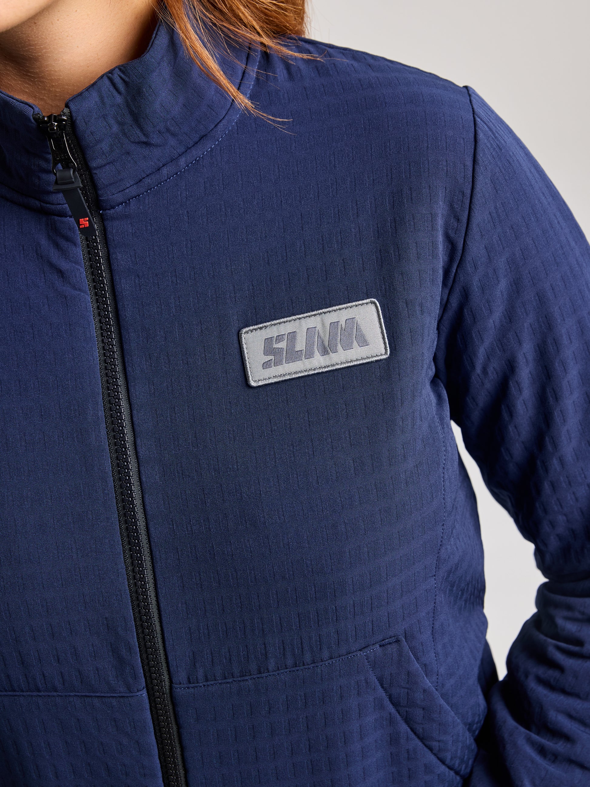 A close-up showcases someone wearing the SLAM MINNIE BEAR JACKET, crafted from recycled waterproof fabric in a quilted blue style. The jacket is adorned with a logo patch on the chest and features a front zipper along with a high collar, embodying the ideal combination of style and functionality inspired by nautical sportswear. Only the upper torso and part of the right arm are visible.