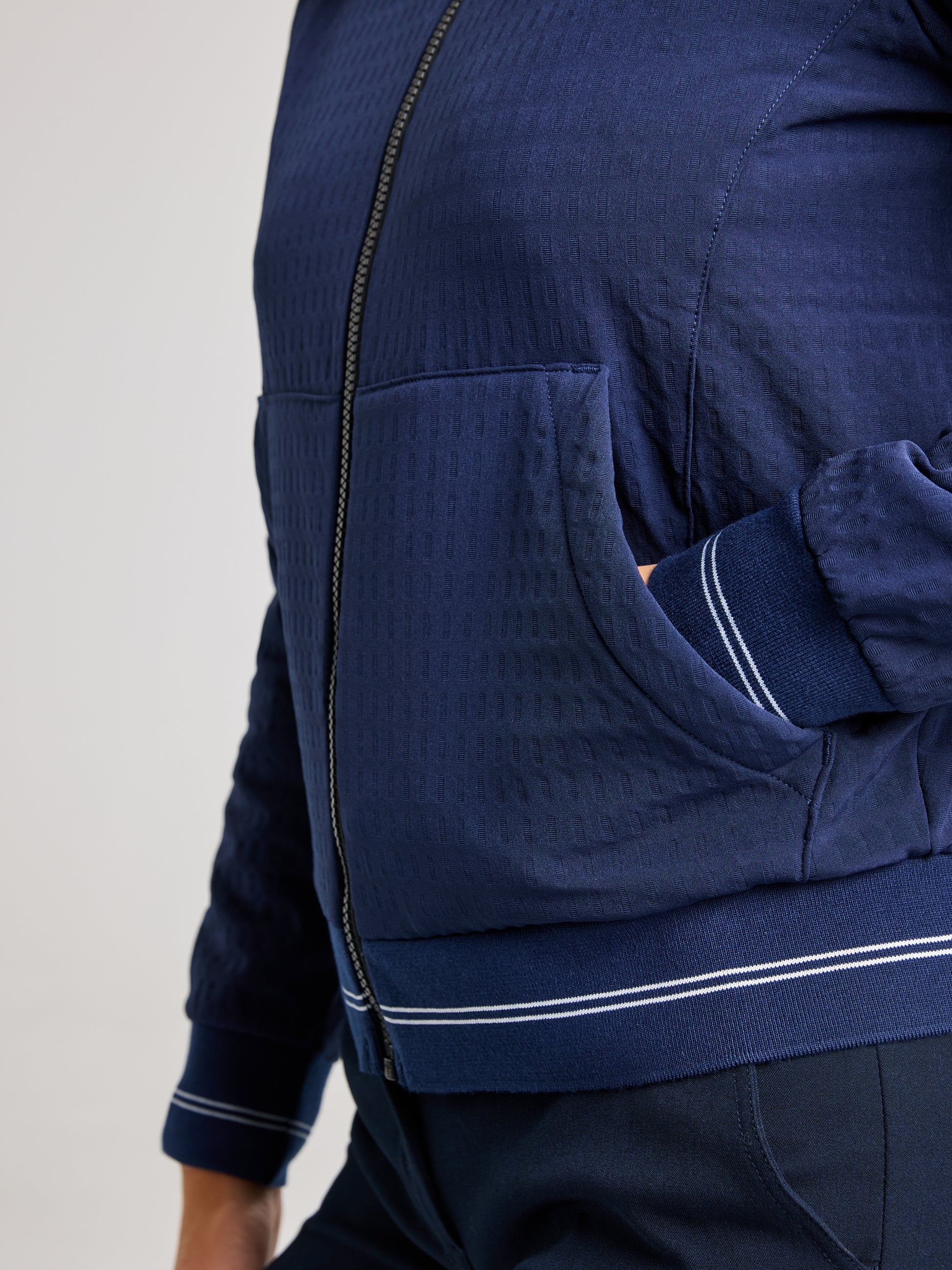 The individual is wearing the SLAM MINNIE BEAR JACKET, made from textured dark blue recycled waterproof fabric. Their hand is tucked into a front pocket, and the look is finished with ribbed cuffs and a hem featuring white stripes. Paired with this nautical sportswear style are matching dark blue pants.