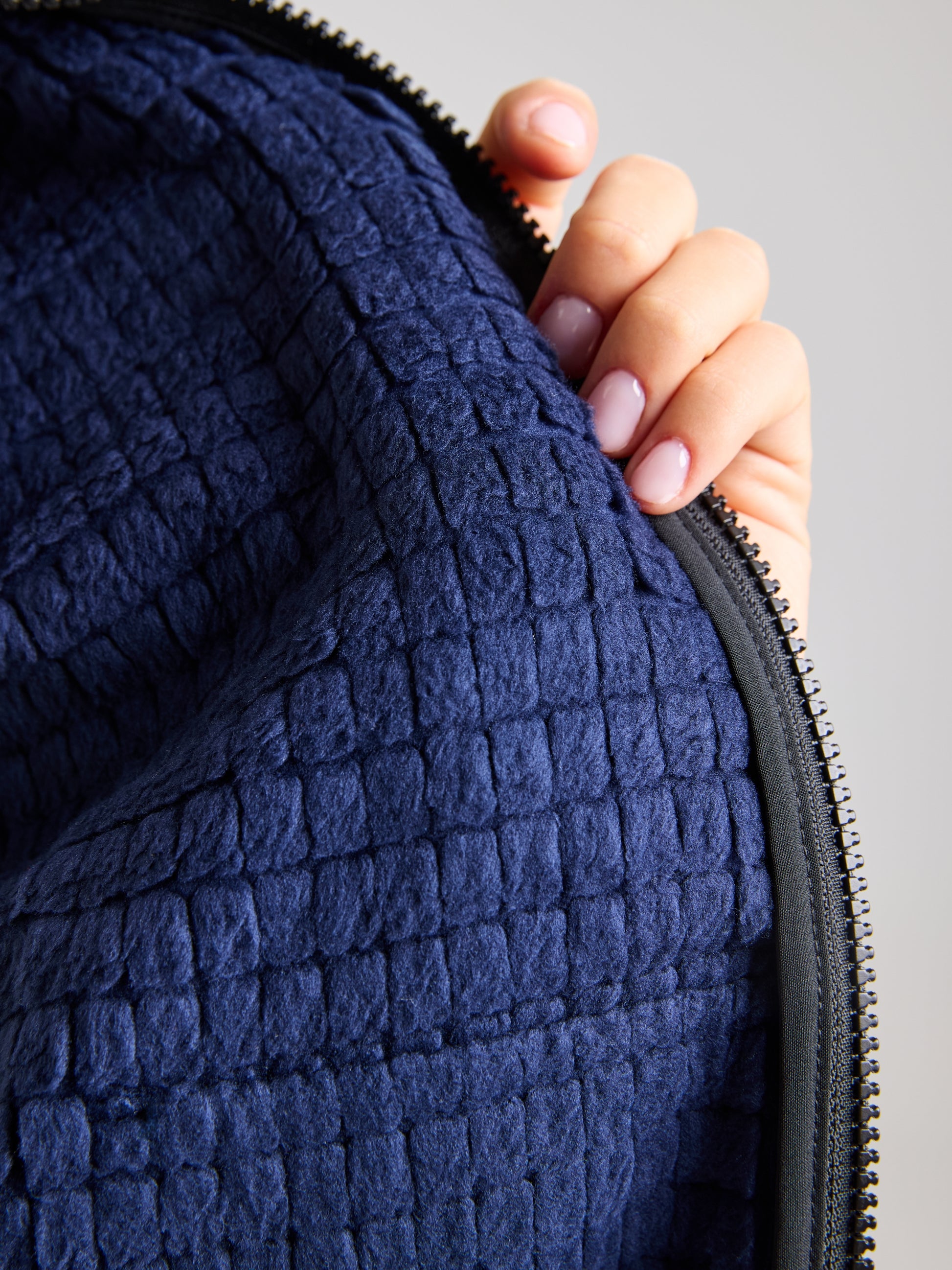 A person with light pink nails holds the MINNIE BEAR JACKET by SLAM, showcasing its blue textured fabric that evokes a nautical sportswear vibe. The jacket features a grid-like pattern and is partially enclosed by a black zipper, all set against a neutral gray background that highlights the elegance of its design.