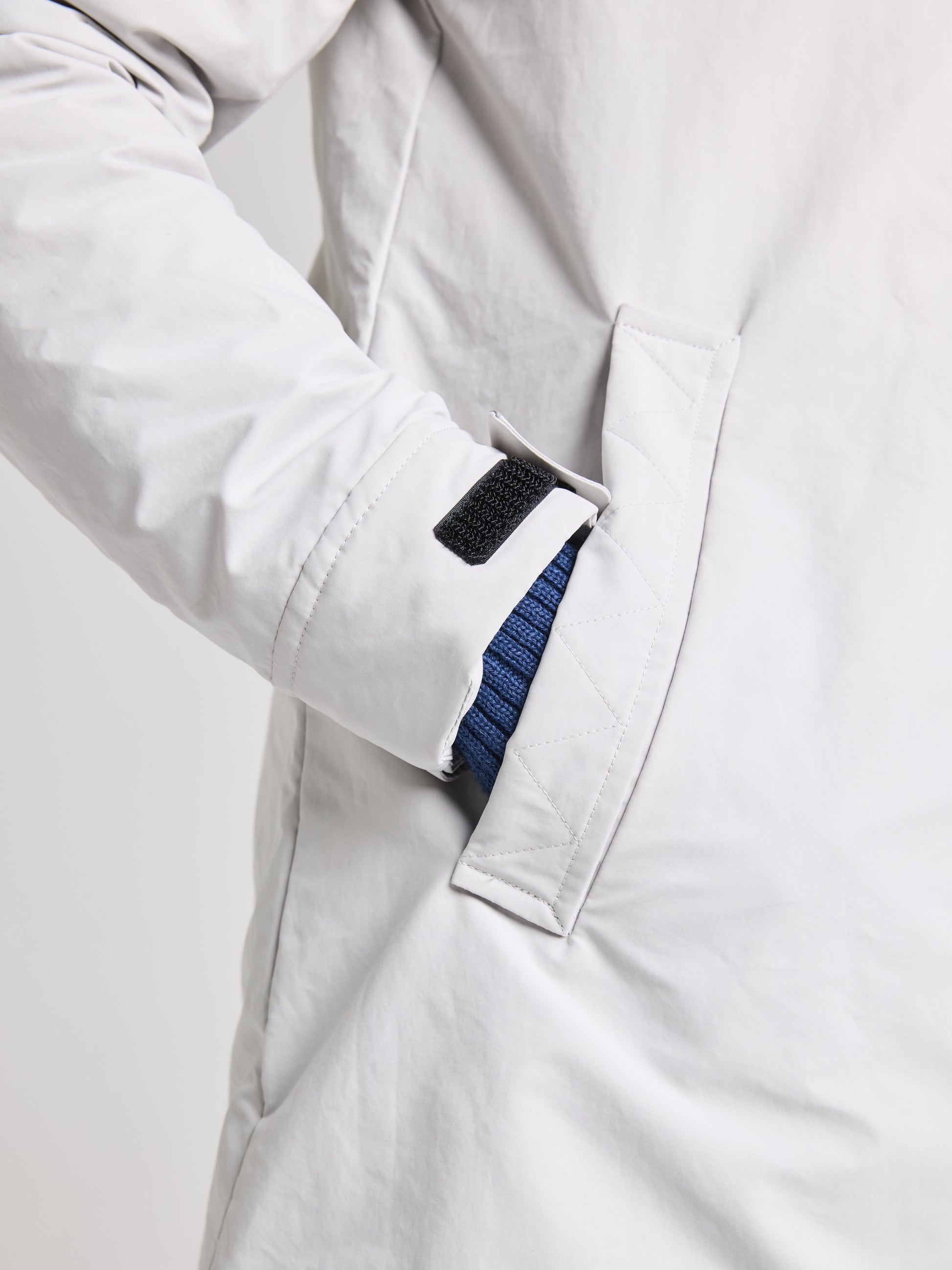 A person is seen wearing the SLAM URBAN WOMAN COAT in gray, with their hand tucked into a side pocket. The hand is shielded by a blue cuff, suggesting additional layers underneath this waterproof coat. It boasts quilted stitching and a Velcro strap near the wrist, making it ideal for cool city escapades.