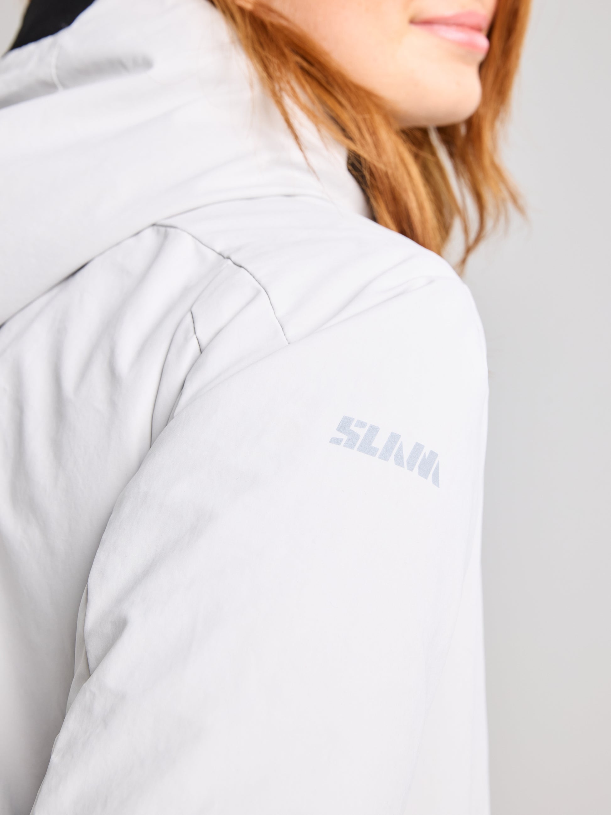 A close-up shot captures a person wearing the URBAN WOMAN COAT by SLAM, in light grey, with the logo prominently displayed on the sleeve. The stylish jacket, featuring hand-warming pockets, complements their shoulder-length reddish-brown hair as they face away from the camera, showing only part of their face and shoulder.