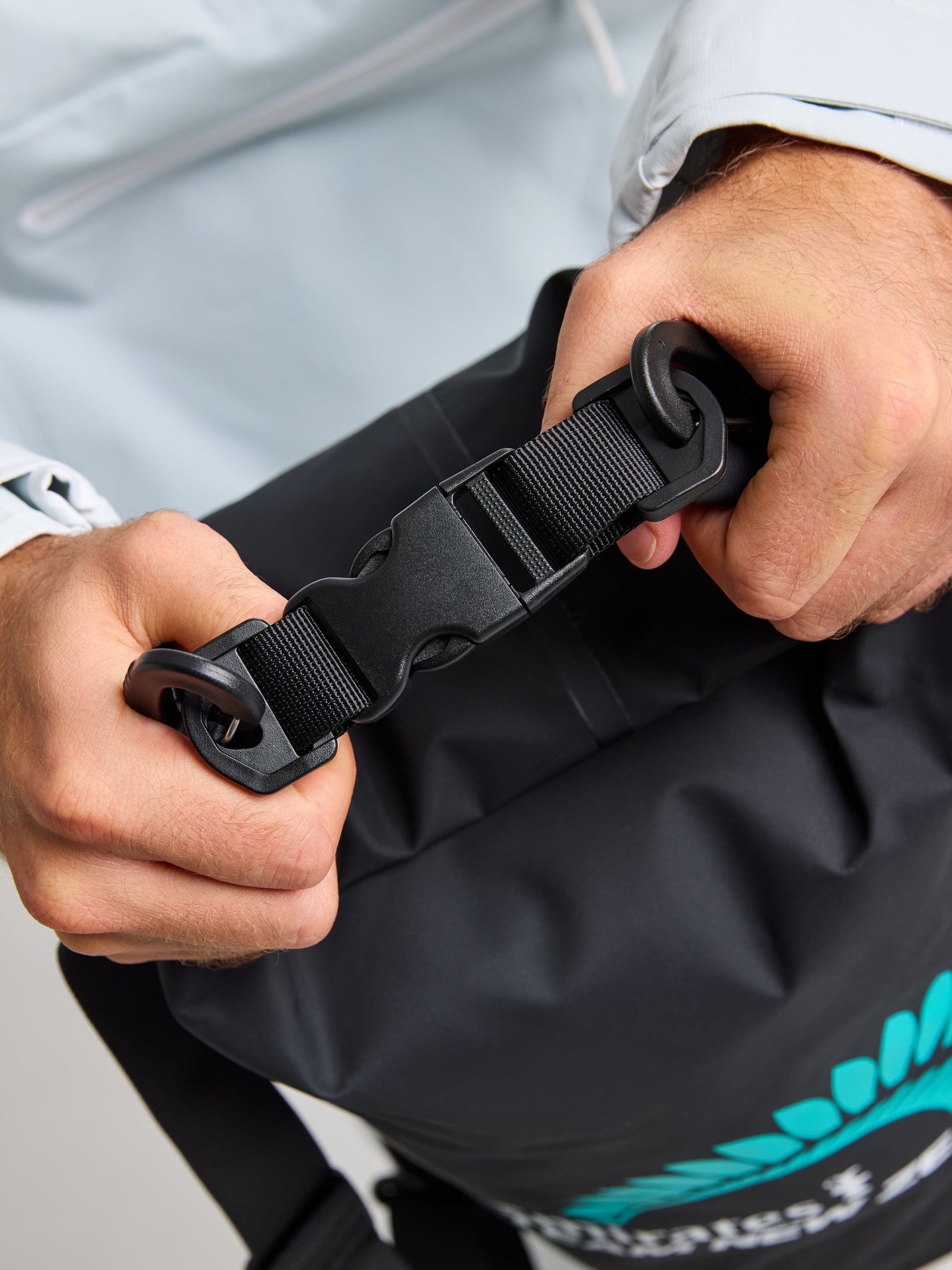 A person, dressed in a white jacket, is securing the buckle on a black ETNZ WR Sailor Bag S by SLAM. Made from water-repellent technical fabric, the unisex beach bag showcases a turquoise graphic design that is partially visible. The person's hands are adjusting the strap to ensure a secure closure.