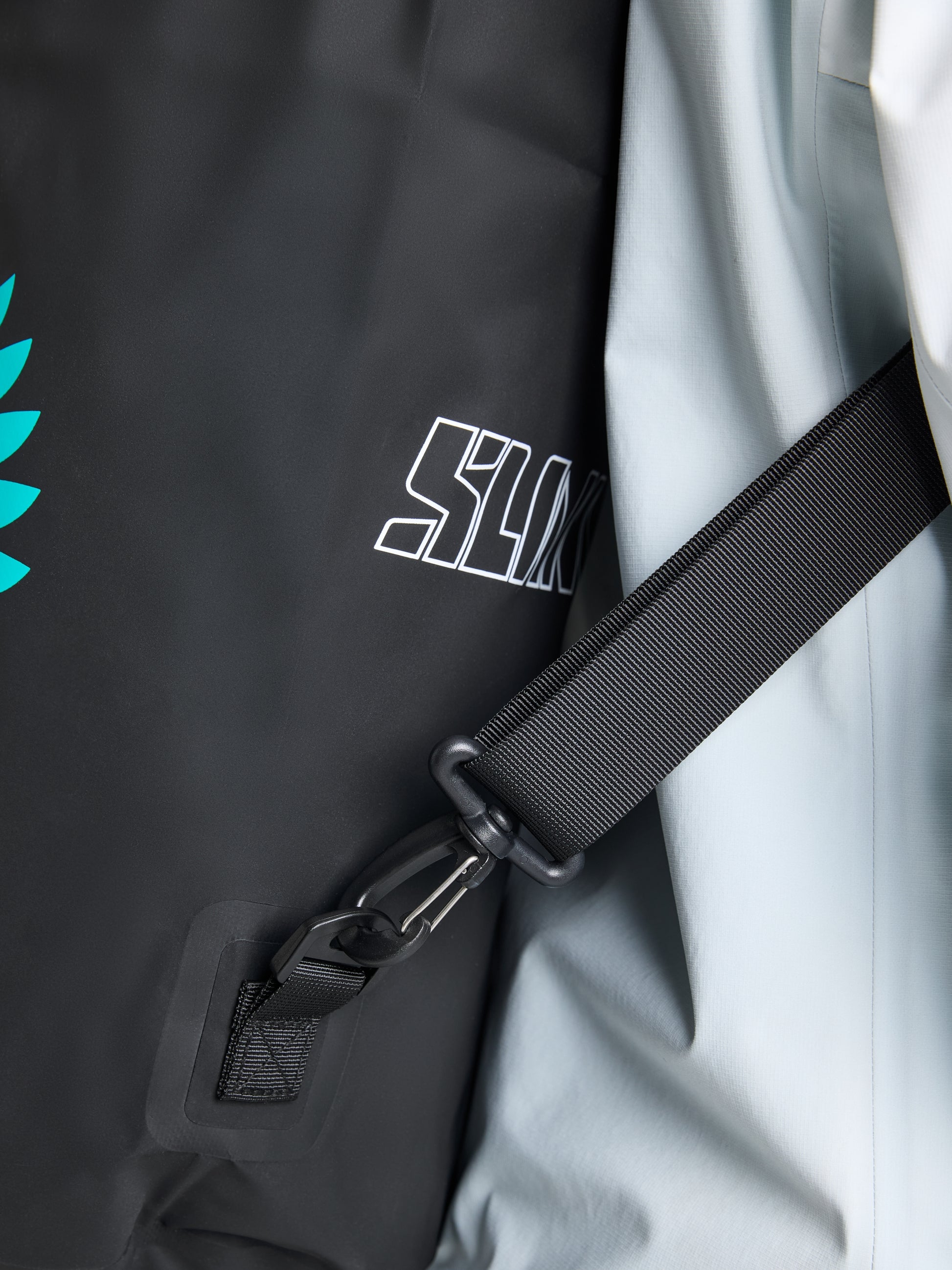 Close-up of the ETNZ WR Sailor Bag S by SLAM, a black and white backpack made from technical fabric. It features a black adjustable strap and buckle, adorned with a turquoise leaf design and partial white text. The smooth, slightly shiny white section highlights its water-repellent quality, making it perfect for any adventure.